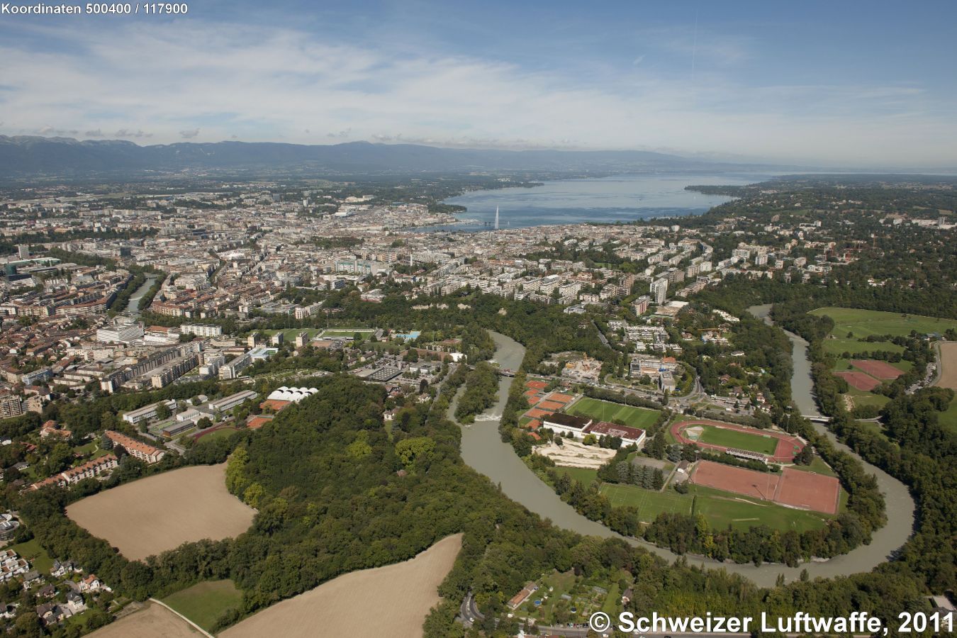 Genf / Genève, Blick gegen Norden zur Stadt mit dem 'Jet d'eau'. - Rhoneschlaufe im Vordergrund: Sportanlagen 'Le Bout-du-Monde' (Stade d'athlétisme du Bout du Monde); Position: 2'500'992.28, 1'115'140.67 - Abgeerntete Felder: 'Sous Pinchat'. - Bildmitte links der Brücke über die Rhone: Stadtteil Fontenette-Gevril.